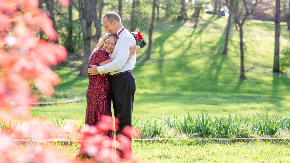 Spring seasonal elopement showing bride in scarlet dress and groom in red bow tie standing and bugging each other with pink treee buds in the foregound.