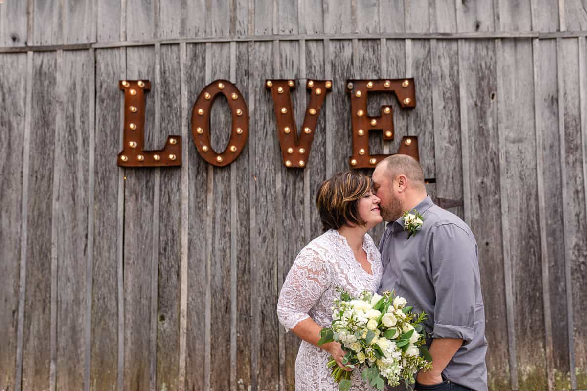 couple kiss next to barn with huge sign in lights reading LOVE