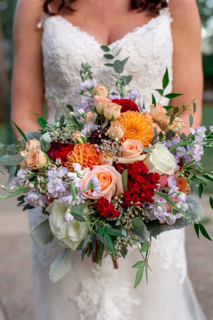 A bride holds a bouquet floral design using red, orange and purple flowers with greenery.