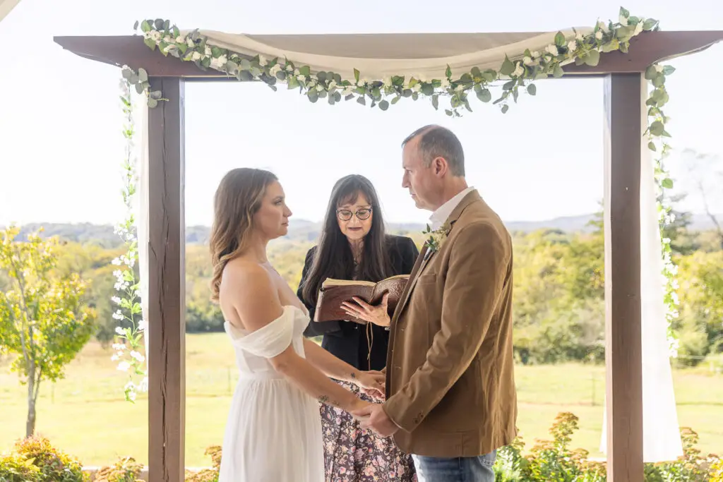 A bride and groom share their wedding vows on a Tennessee farm, standing under a floral draped arch.